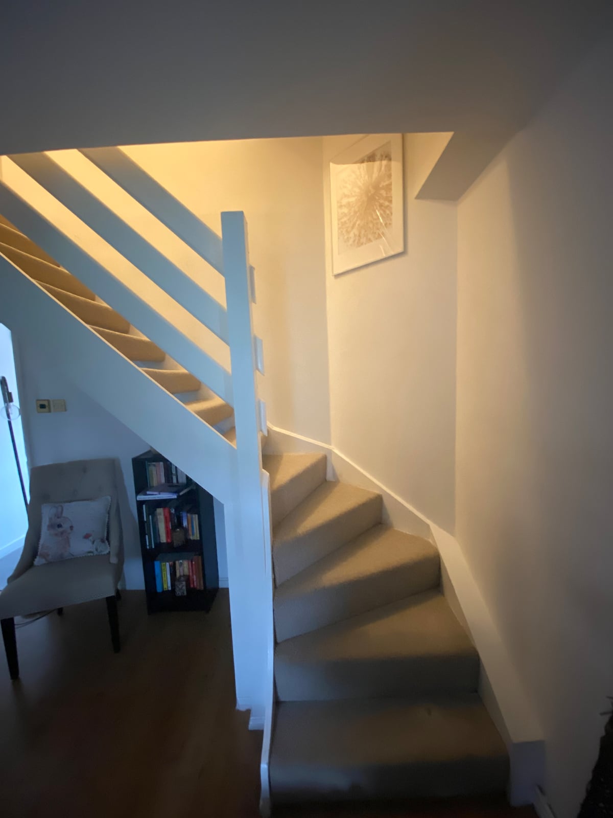 A softly lit staircase ascends, bordered by a white railing. A chair is positioned against the wall, with a small bookshelf displaying various titles nearby. A framed art piece is affixed to the wall above the stairs, adding a touch of decor to the space.