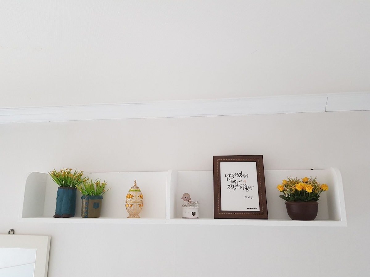 A white shelf mounted on a light-colored wall is displayed. It holds a variety of decorative items, including two small potted plants, a ceramic vase, a framed print with text, and a decorative figure. The arrangement adds a subtle touch of decor to the space.