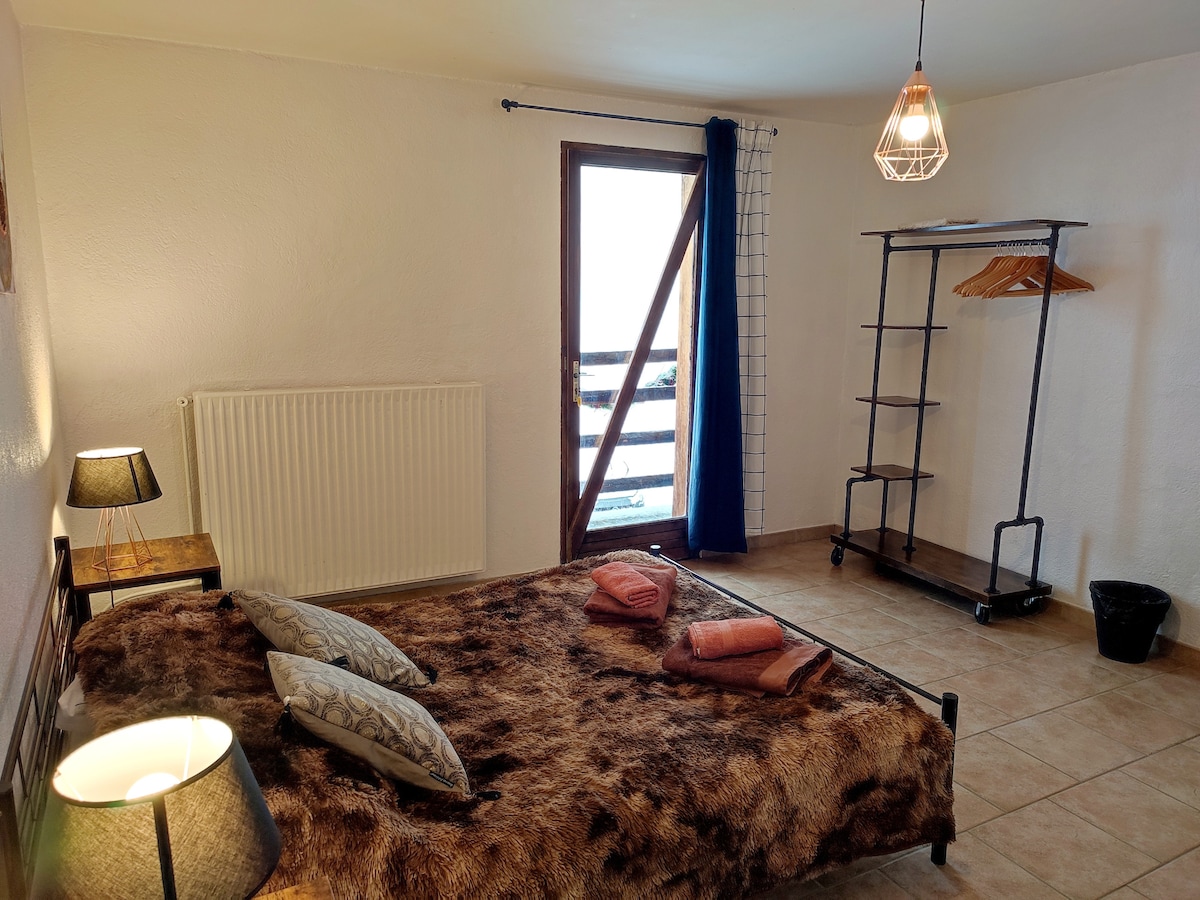 A spacious sleeping area features a fur-textured bedspread and several decorative cushions. Natural light filters through the window, which is framed by minimal curtains. A clothing rack stands against the wall, accompanied by a small wastebasket and two bedside lamps providing soft illumination.