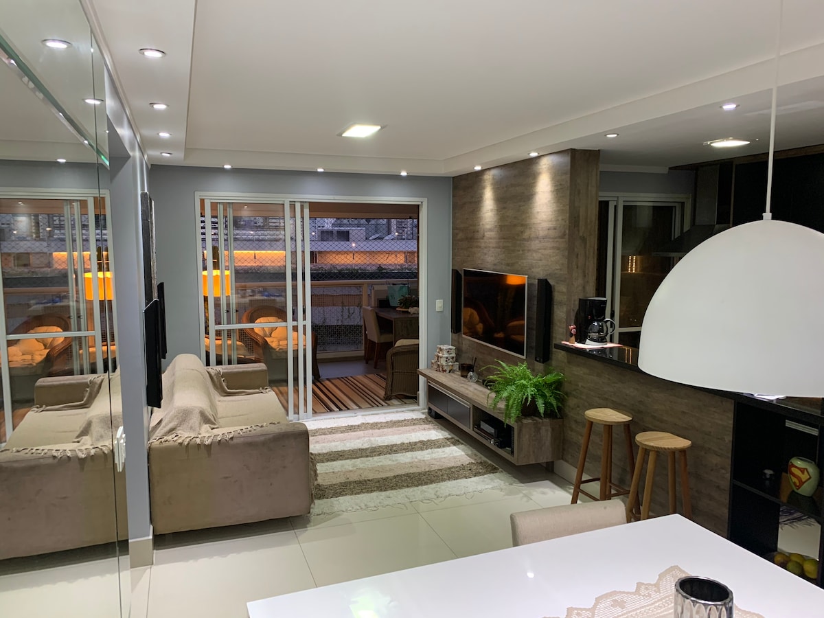 A modern living area is shown, featuring a comfortable couch and a TV mounted on a textured wall. Natural light enters through large sliding doors, illuminating the space. A dining area and kitchen island with stools are visible, adding to the open layout.