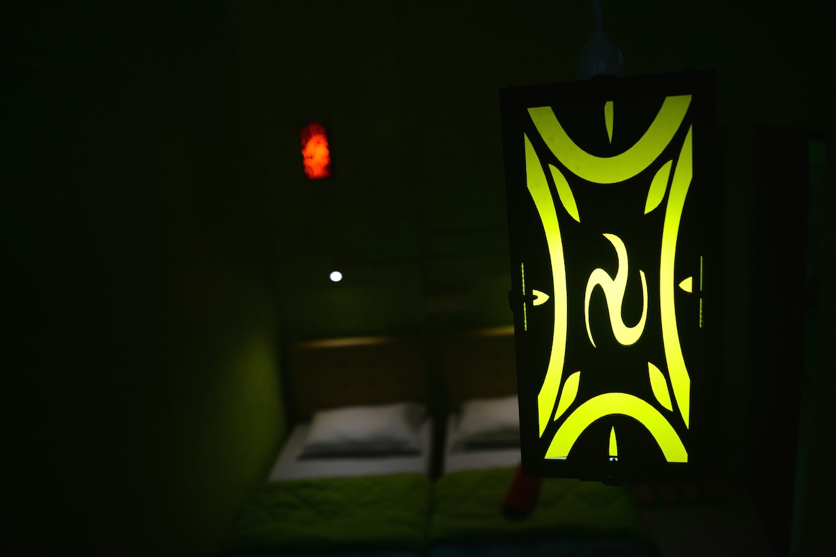 An illuminated green pendant light casts a warm glow over the sleeping area. Two spacious beds are partially visible in the background, featuring crisp white bedding. Soft lighting creates a calm ambiance, enhancing the overall tranquility of the space.