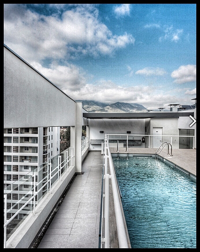 A rooftop swimming pool is surrounded by a white deck and glass barriers, offering views of distant mountains. The pool features clear blue water, with a set of stairs leading into it. The sky is partly cloudy, enhancing the open atmosphere.