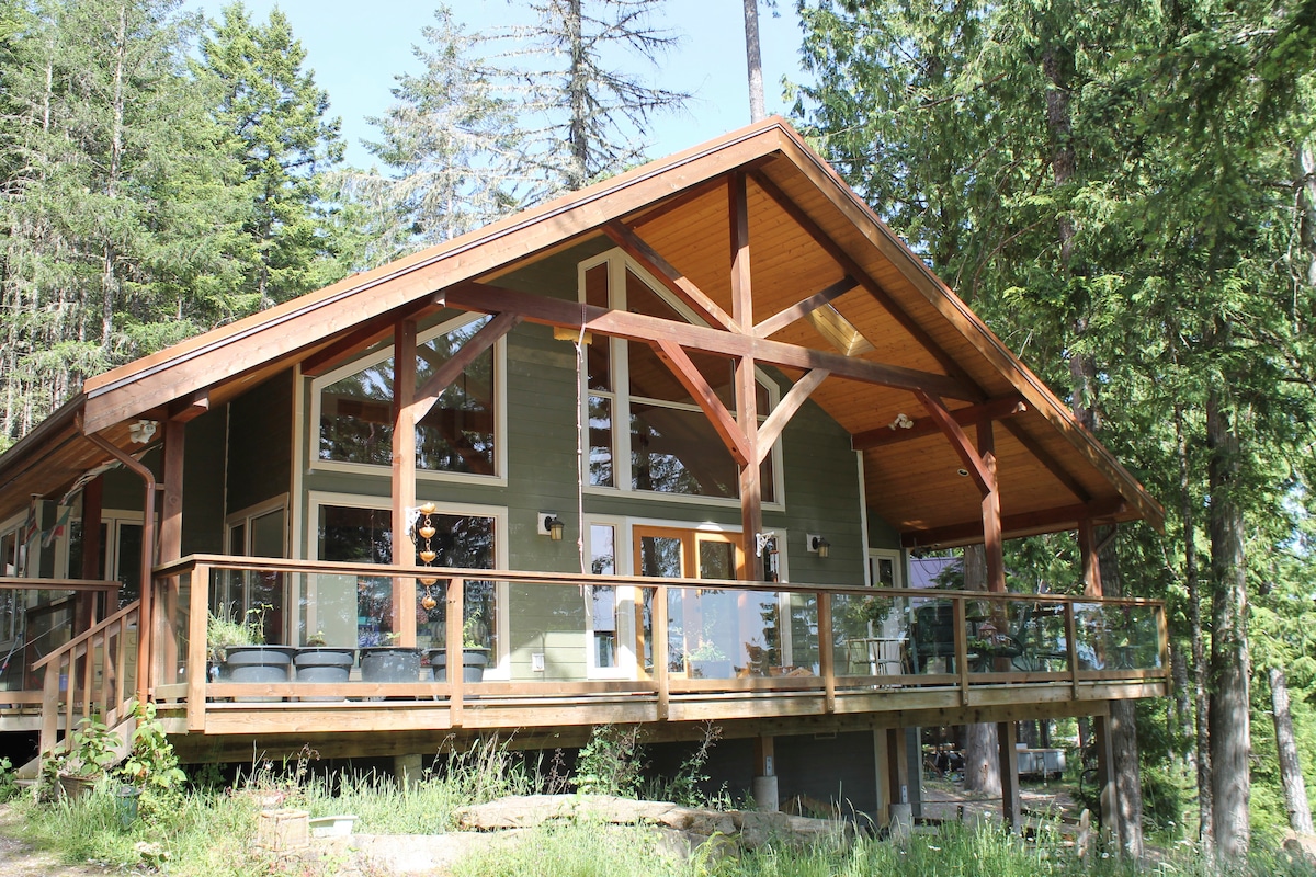 The modern chalet features a spacious deck with a glass railing and wooden beams that highlight its architectural design. Surrounded by tall trees, large windows provide ample natural light and views of the outdoor greenery.