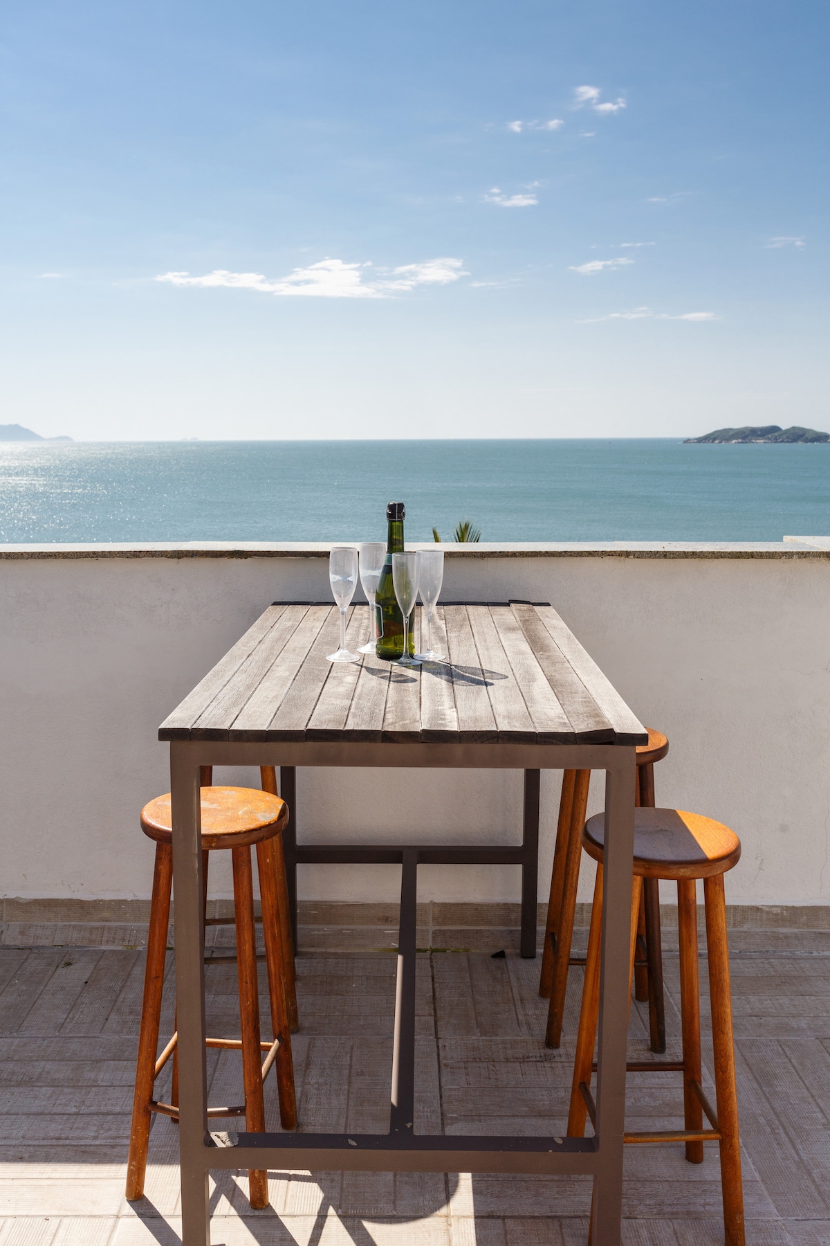 A rooftop terrace table is set with two clear glasses and a bottle, overlooking a scenic ocean view. Two wooden stools are positioned at the table, and the sky is clear, reflecting the tranquil atmosphere. The coastal landscape stretches into the distance.