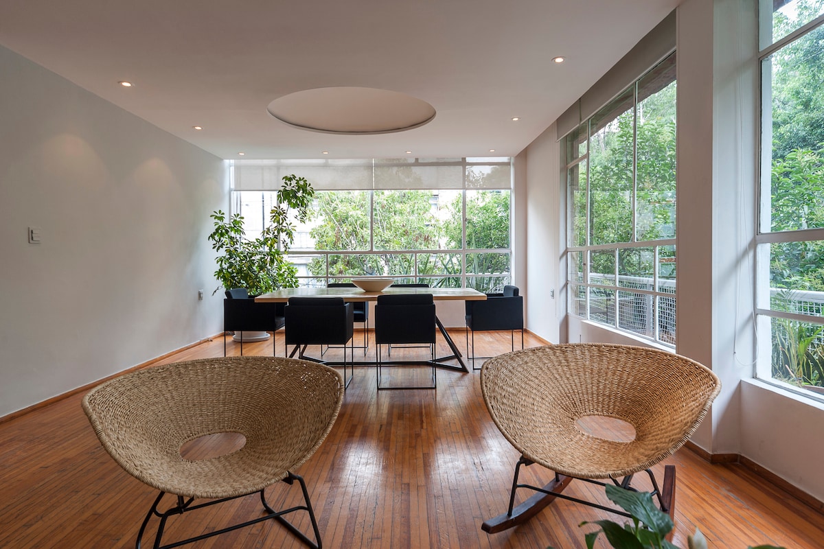 A spacious living area features large windows that invite natural light and offer views of lush greenery. Two distinctive basket chairs are positioned on wooden floors, while a contemporary dining table with black chairs sits in the background, highlighted by a circular ceiling detail.