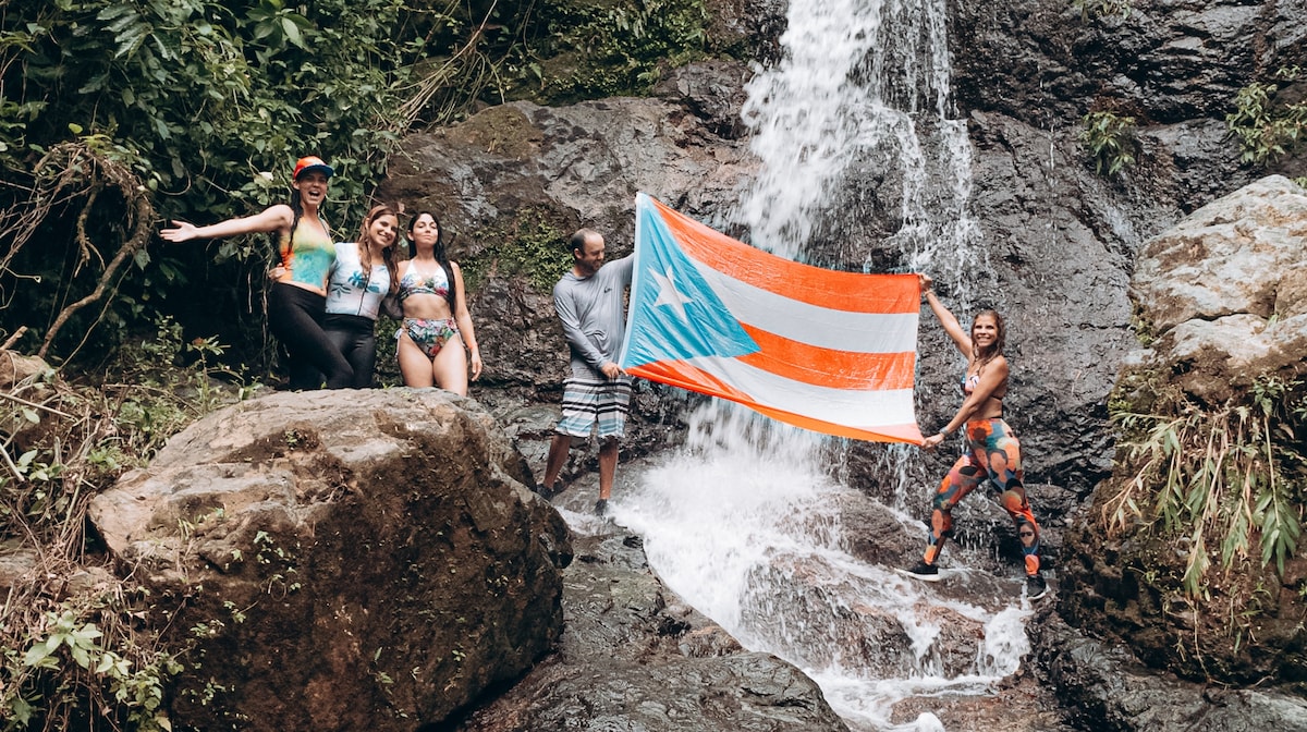 A group of individuals stands near a cascading waterfall, showcasing vibrant flora in the surrounding area. One person holds a Puerto Rican flag, while the others are dressed in swimwear. Large rocks create natural seating near the water's edge.