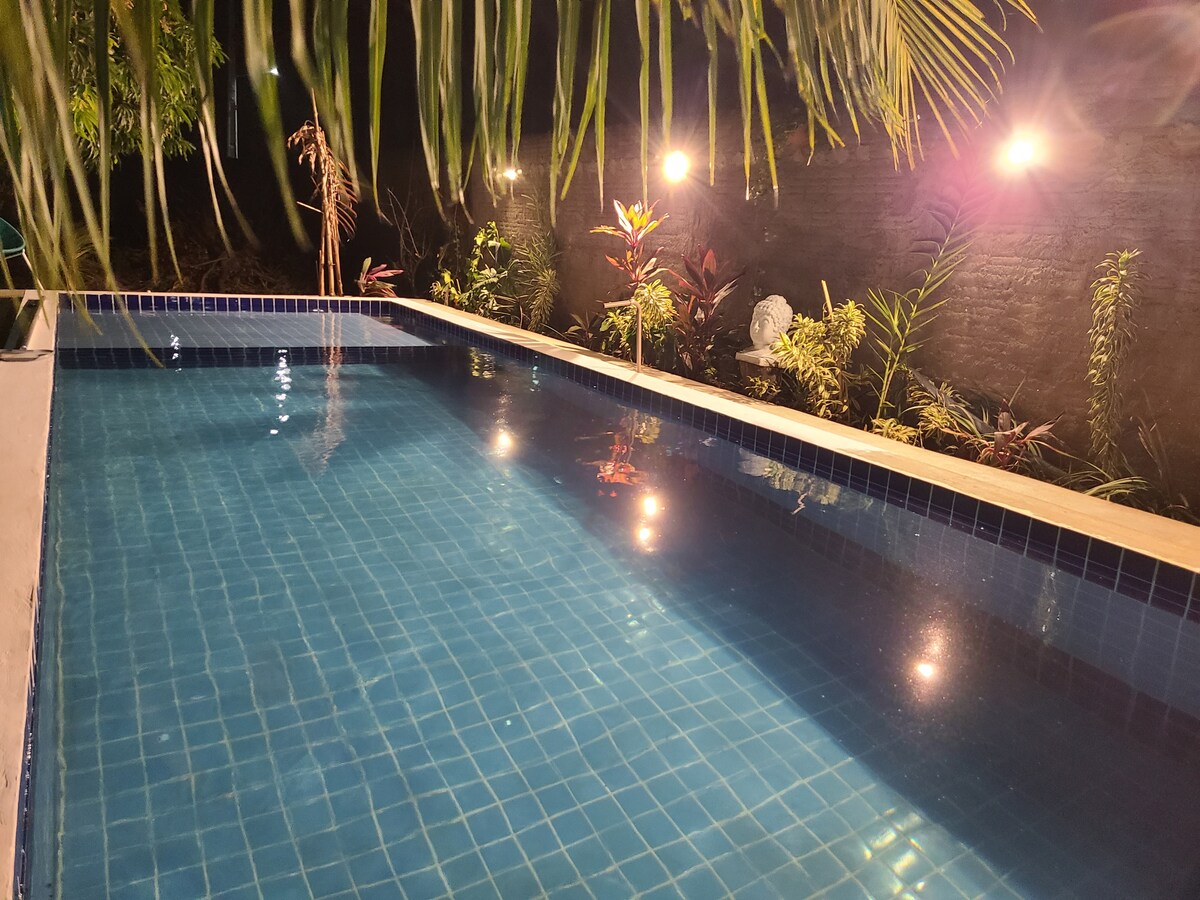A serene swimming pool is nestled among lush greenery, with soft lights creating a calming ambiance. The water reflects the surrounding plants and illuminated surfaces, inviting relaxation and connection with nature.