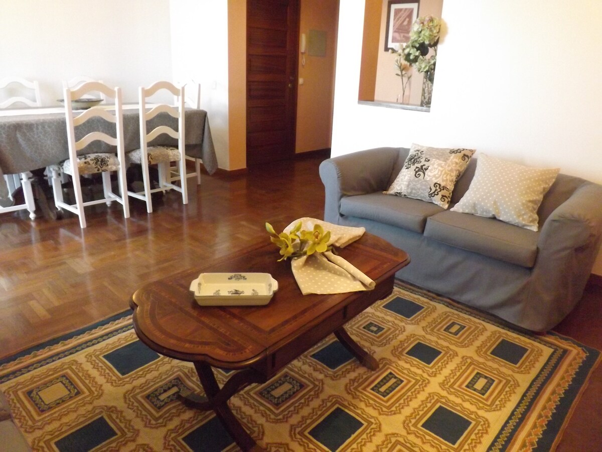 A living area features a spacious gray sofa complemented by decorative cushions. A wooden coffee table with a floral arrangement sits in front. A patterned rug covers the floor. In the background, a dining area with a large table and white chairs is visible.