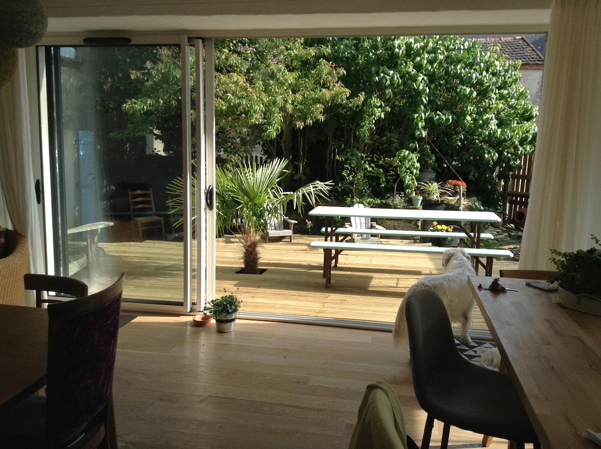 A spacious indoor area opens to a wooden deck through large glass sliding doors. A long picnic table is set on the deck, surrounded by various plants and greenery, creating a natural atmosphere. Soft light filters through sheer curtains, enhancing the warm environment.