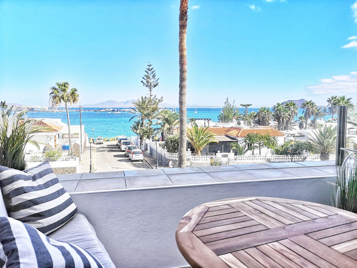 A terrace offers views of the sea and surrounding palm trees, with a wooden table and a cushioned seating area visible. The clear blue sky complements the vibrant colors of the beach area in the distance, creating a serene outdoor space.