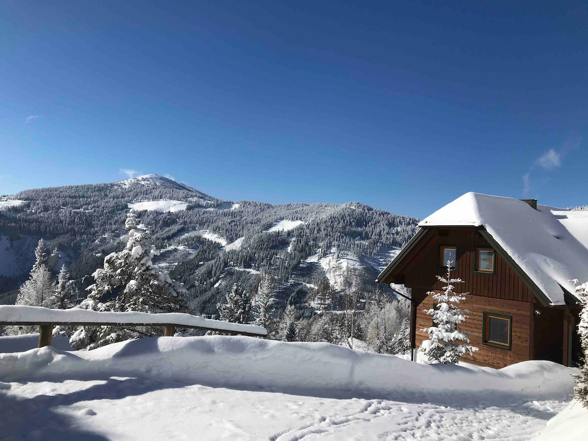 A wooden chalet is positioned next to gently sloping snow-covered hills, with a clear blue sky above. Surrounding fir trees are dusted with snow, creating a serene winter landscape. A wooden railing frames the view, enhancing the natural beauty of the surroundings.