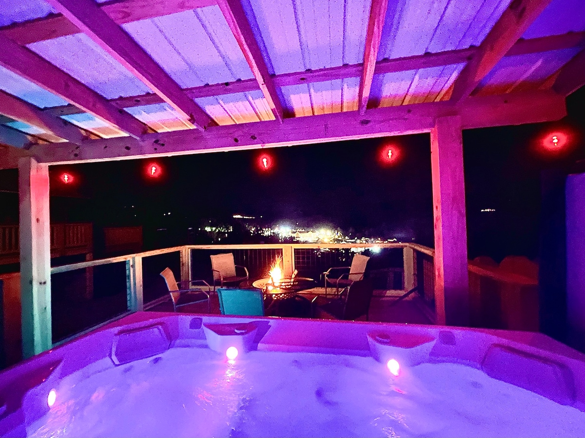 A large hot tub, illuminated with soft purple lights, is positioned under a wooden pergola. Surrounding the tub, simple outdoor furniture offers a cozy seating area. In the background, hillside views are complemented by warm lighting from nearby buildings, creating a serene nighttime atmosphere.