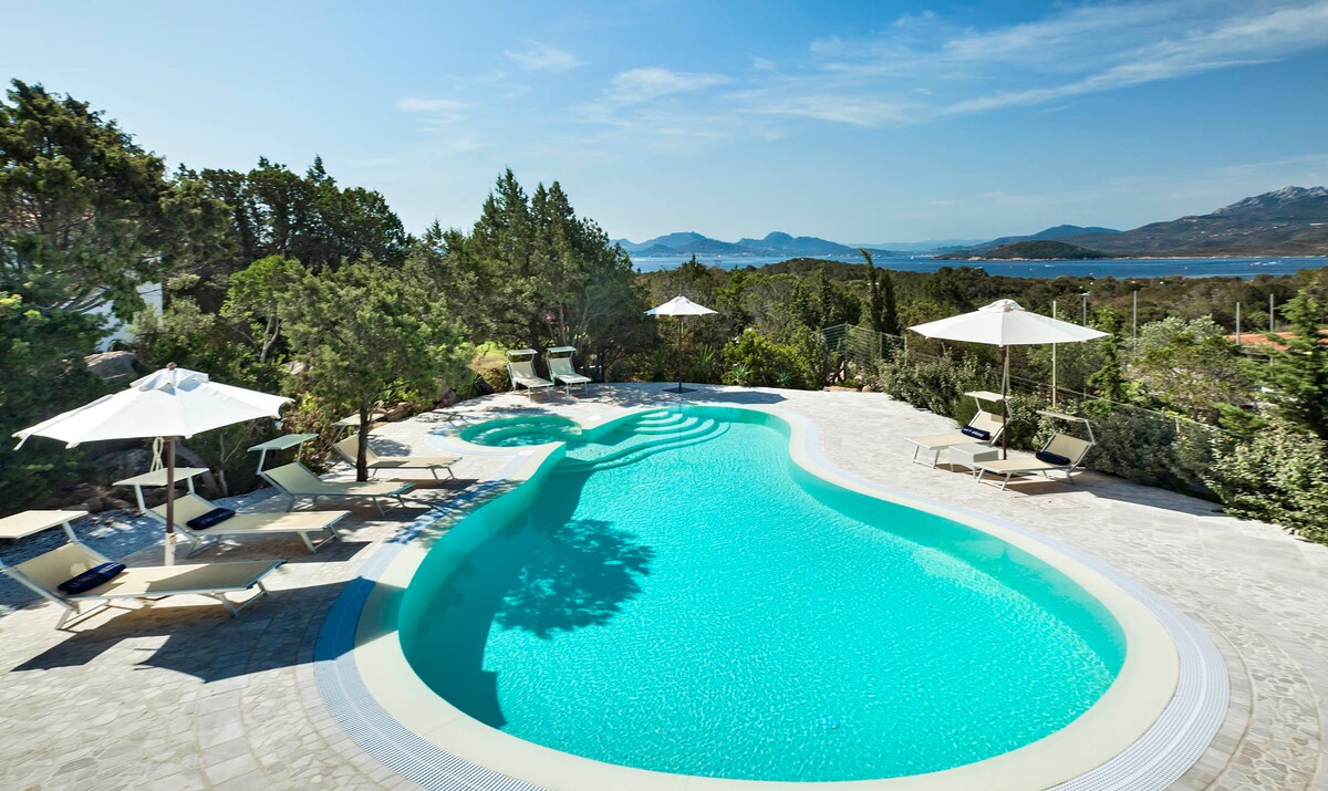 A serene swimming pool is outlined by natural stone, with clear turquoise water gently reflecting the sky. Sun loungers are positioned around the pool, and white umbrellas provide shade, surrounded by lush greenery and distant views of the bay.
