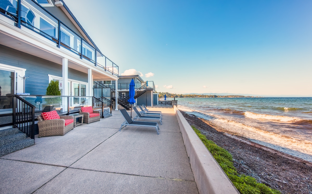 An outdoor patio area features comfortable seating and sun loungers arranged along the oceanfront. The smooth concrete surface leads to a pebbled beach, with waves gently lapping the shore. An inviting blue umbrella provides shade, complementing the vibrant red cushions on the chairs.