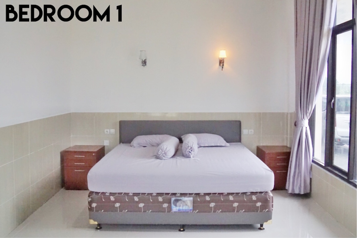 A spacious bedroom features a large bed with light purple bedding, accompanied by two cylindrical pillows. Nightstands with drawers are positioned on either side of the bed. Natural light enters through a large window, with sheer curtains gently framing the view.