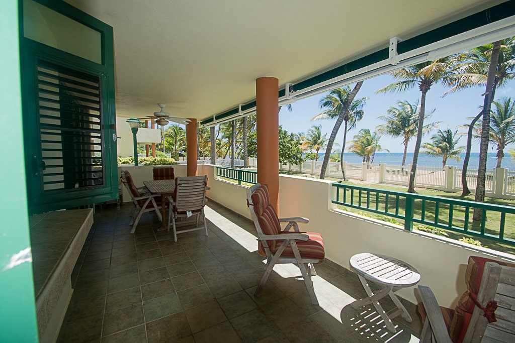 The spacious beachfront veranda showcases a tiled floor and is lined with large windows, allowing natural light and ocean breezes. Comfortable seating options include striped lounge chairs and a small wooden table. Palm trees are visible through the transparent glass, enhancing the coastal ambiance.