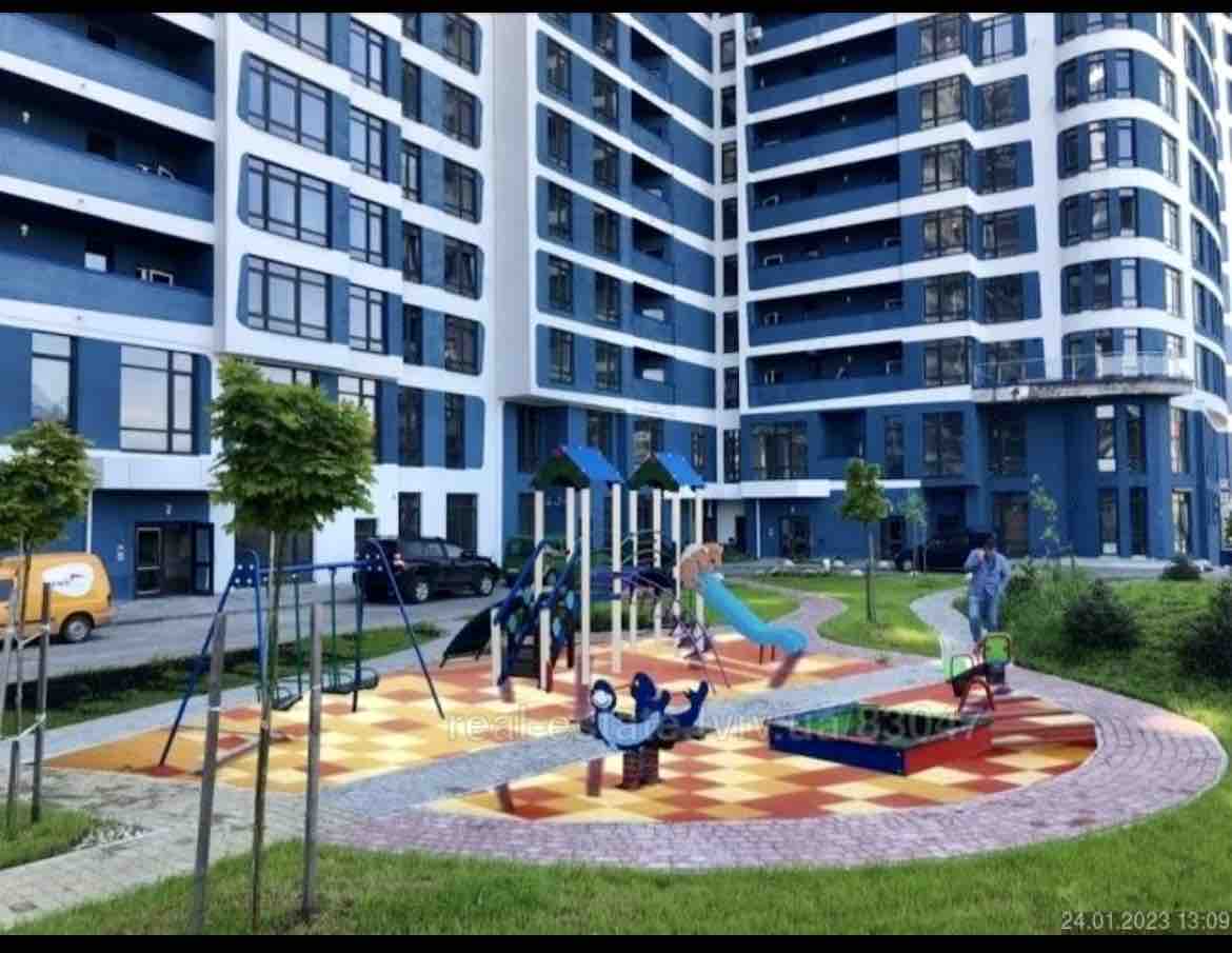 A children's playground is visible, featuring colorful equipment set on a patterned ground. Surrounding greenery includes small trees and landscaped areas. The multi-story building in the background showcases large windows and balconies, creating a vibrant community atmosphere.
