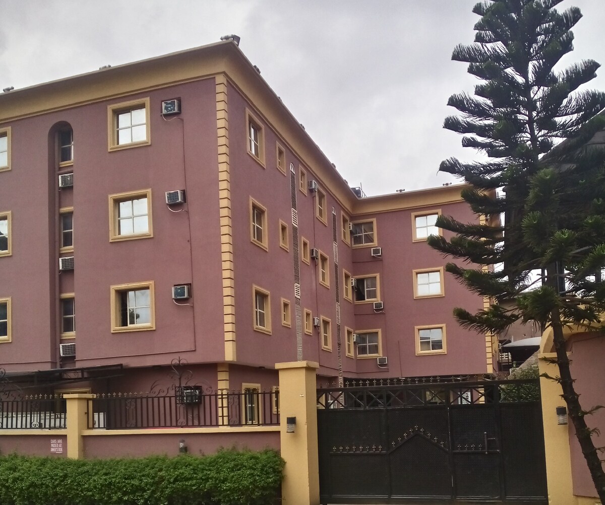 The exterior of the hotel displays a structured, four-story building painted in a warm pink hue with cream-colored trimmings. Multiple air conditioning units are affixed to the walls, and the entrance features a secure gate surrounded by greenery.