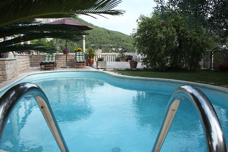 A private pool is surrounded by a well-kept garden, featuring loungers positioned beside it for relaxation. Lush greenery and potted plants enhance the peaceful ambiance. In the background, a terrace is visible, offering scenic views of the nearby hills.