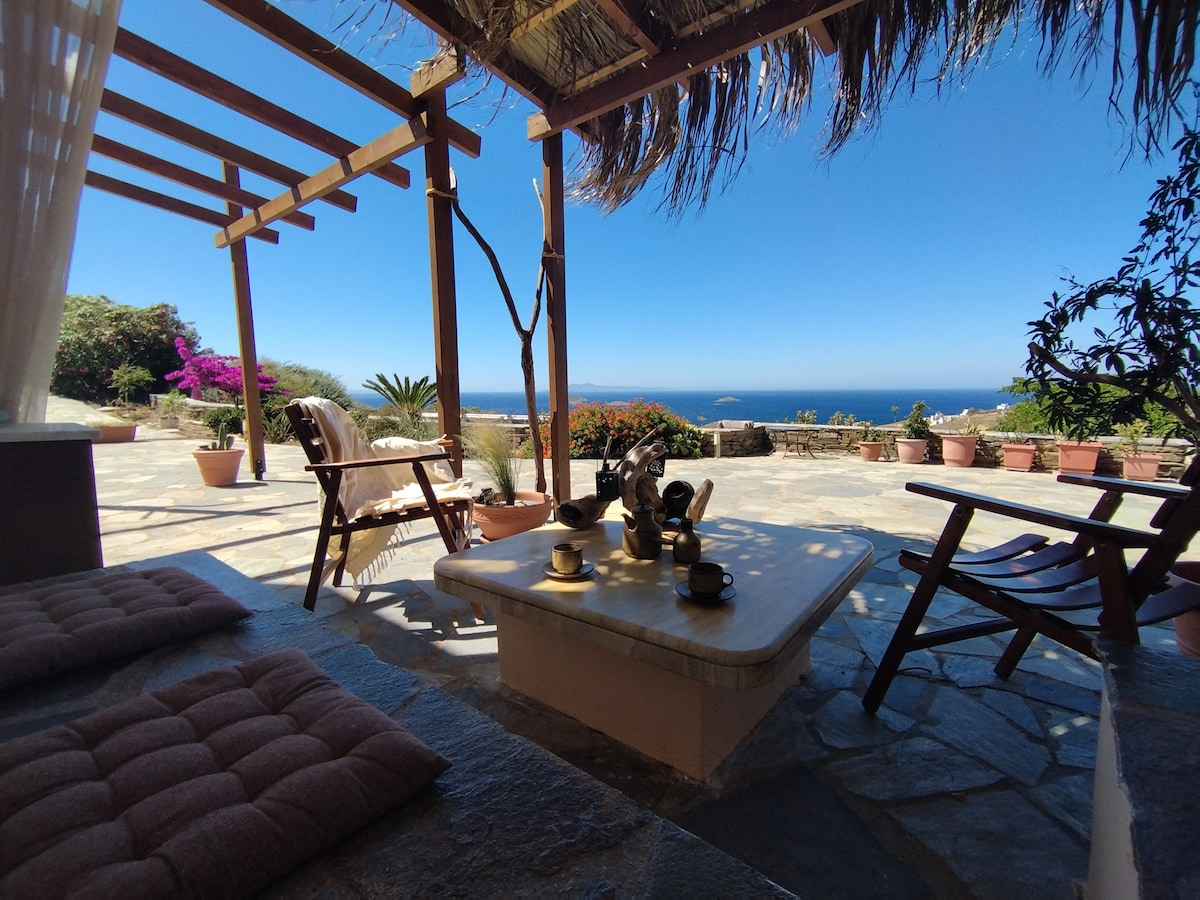 A shaded outdoor area features a stone table set with cups, surrounded by comfortable seating options. Colorful potted plants add greenery, and a stunning view of the Aegean Sea is visible beyond, enhancing the tranquil setting.