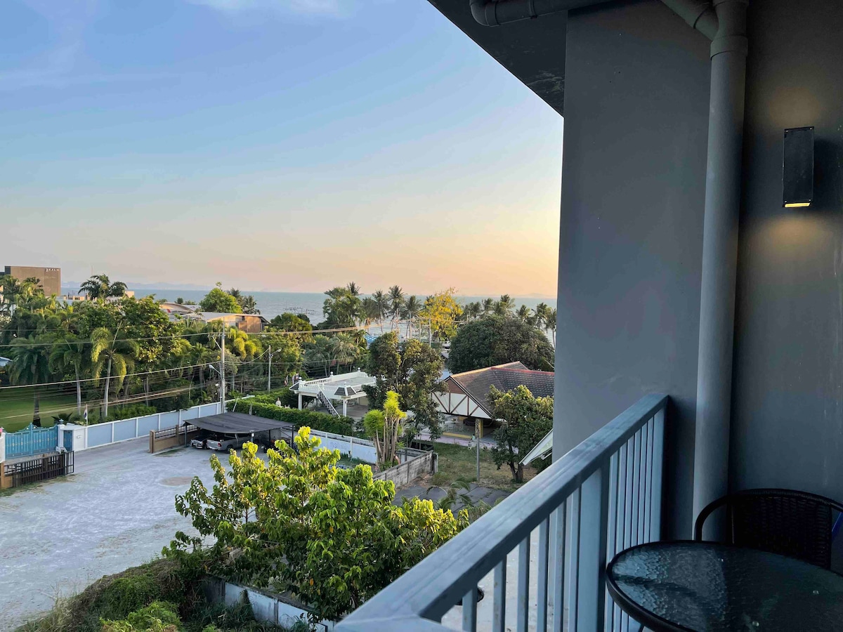 A balcony is visible, featuring a small round table accompanied by a chair. The scene showcases a serene view of palm trees and nearby buildings. The horizon displays soft hues of a sunset, with the sea gently visible in the distance.