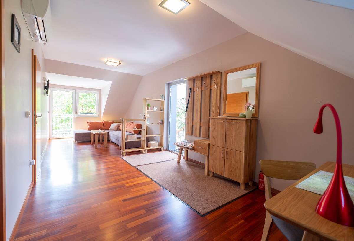 A spacious living area is shown with warm wooden flooring. Natural light streams through the large windows, illuminating a cozy seating area and a brown sofa. An office table and a stylish red lamp complement the simple wooden furnishings, providing a welcoming atmosphere.