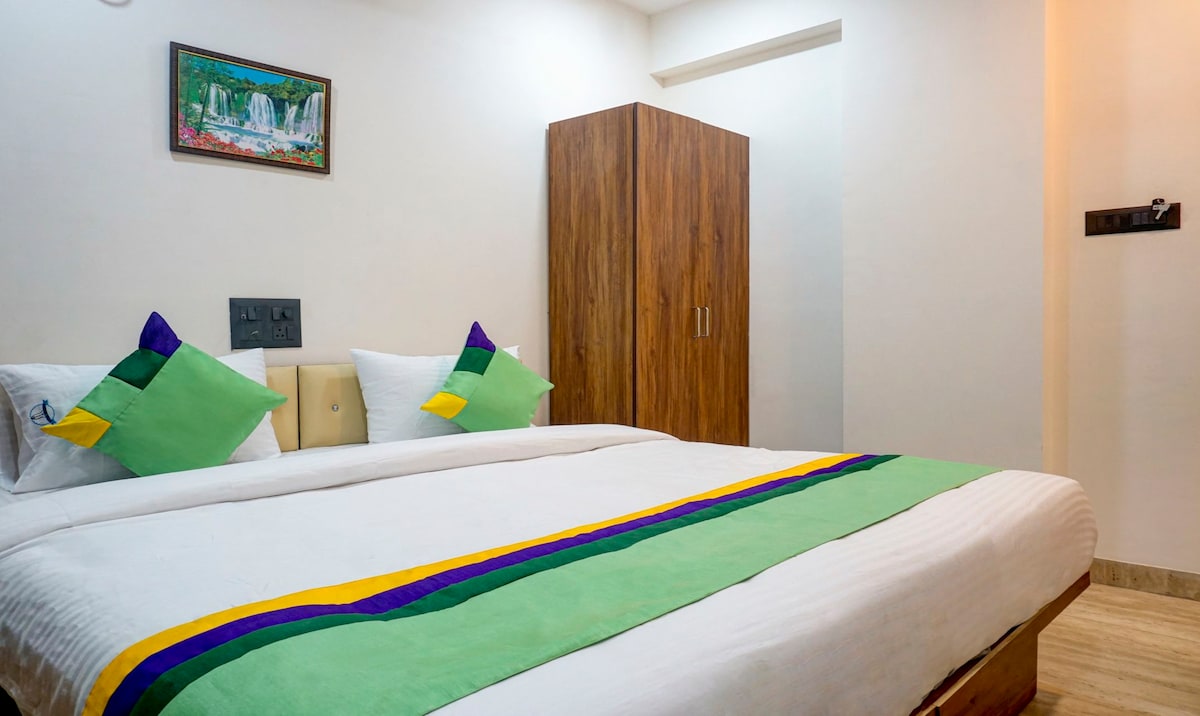A spacious room features a large bed with green and purple accents. Two decorative pillows sit at the head of the bed. A wooden wardrobe is positioned against the wall, and a wall-mounted painting adds a touch of decoration to the space.