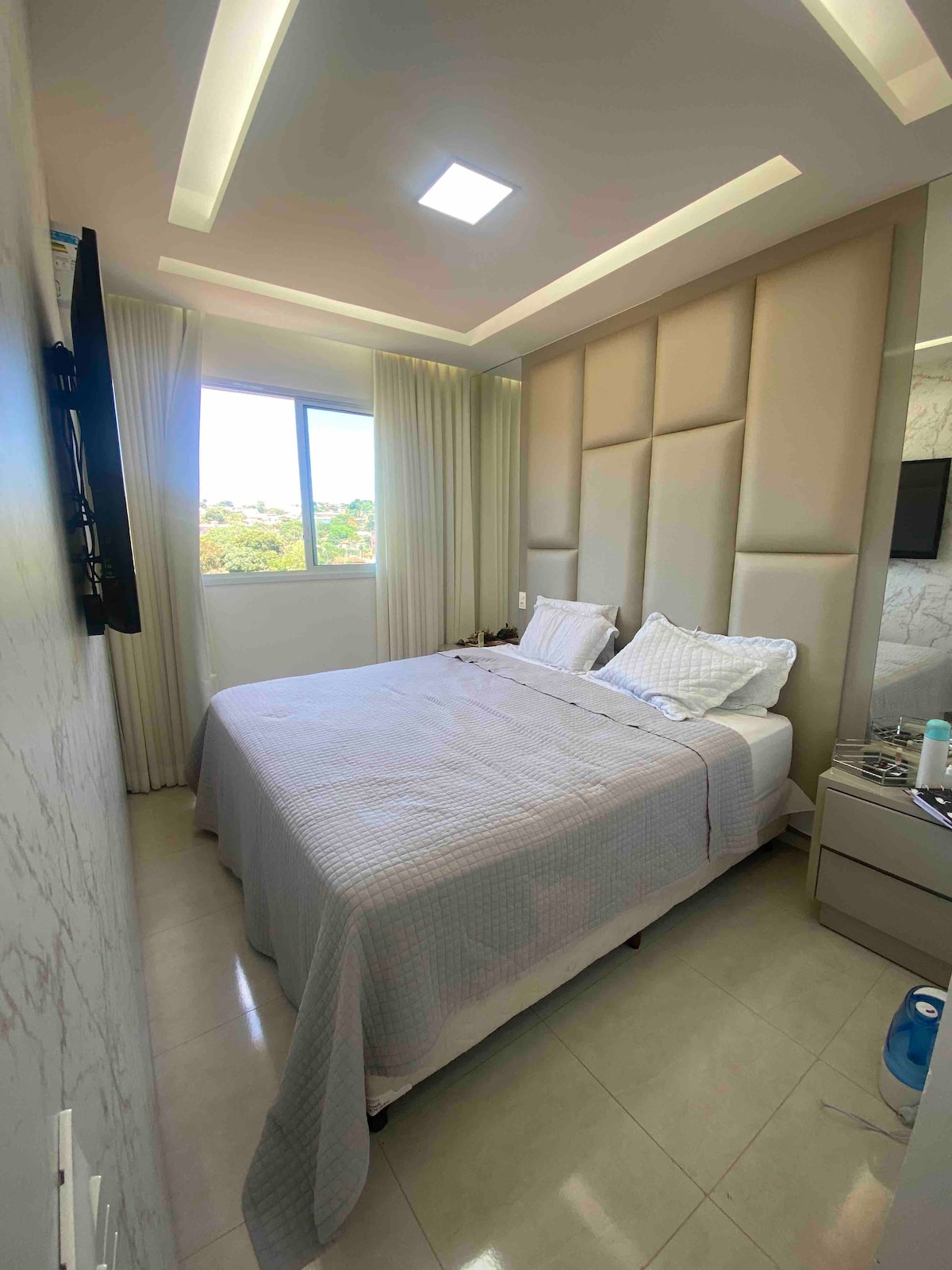 A well-appointed sleeping area features a spacious bed dressed in soft linens. Natural light flows through a large window, which is framed by sheer curtains. A minimalist nightstand sits beside the bed, complemented by an elegant accent wall and modern lighting.