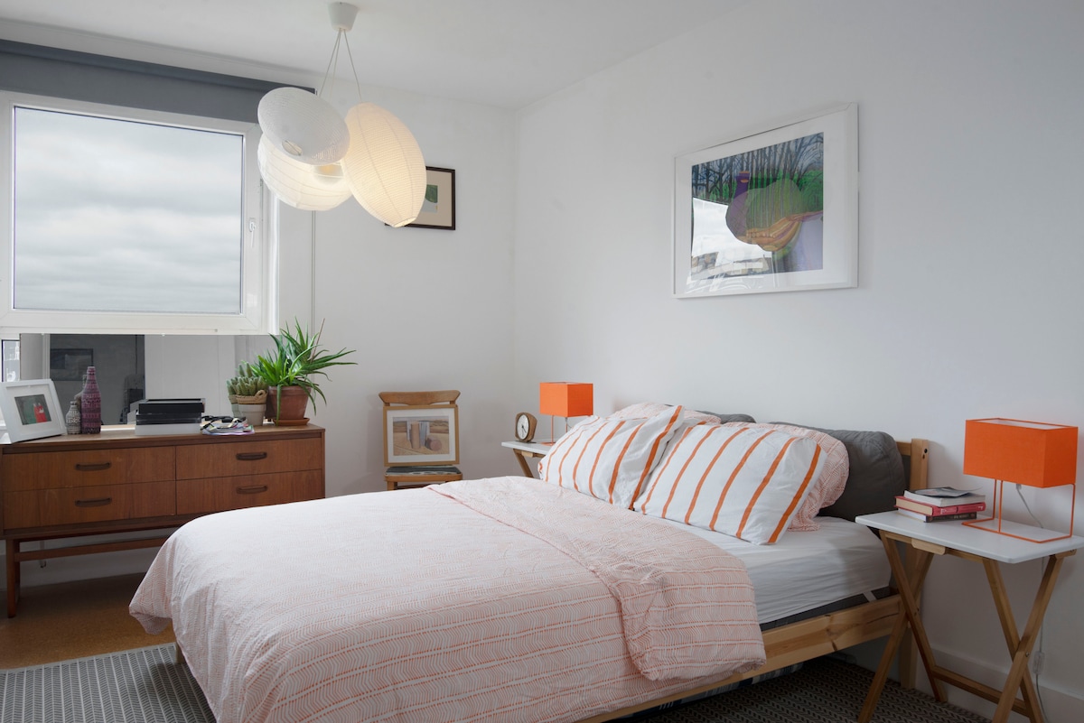 A double bedroom features a spacious bed adorned with a light-colored striped blanket. Bright orange lamps flank the bed, complementing the wooden dresser and framed artwork on the wall. A large window allows natural light to fill the space, with a potted plant adding a touch of greenery.