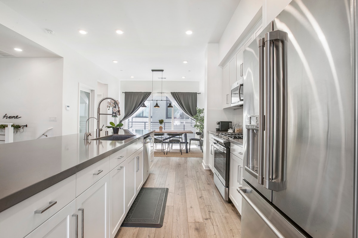 The kitchen features sleek stainless steel appliances and a large island with a contemporary design. Ample storage is provided by white cabinetry. Natural light floods the space through large windows, complemented by elegant drapes. A dining area with a table is visible in the background.