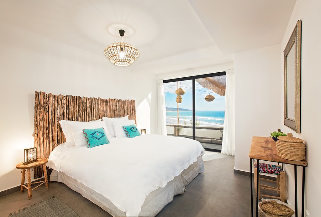 A spacious room is highlighted by a large bed with white linens and decorative blue cushions. Natural wood accents form a striking headboard. A sliding glass door offers direct views of the ocean, while light filters through sheer curtains. A small wooden table complements the decor.