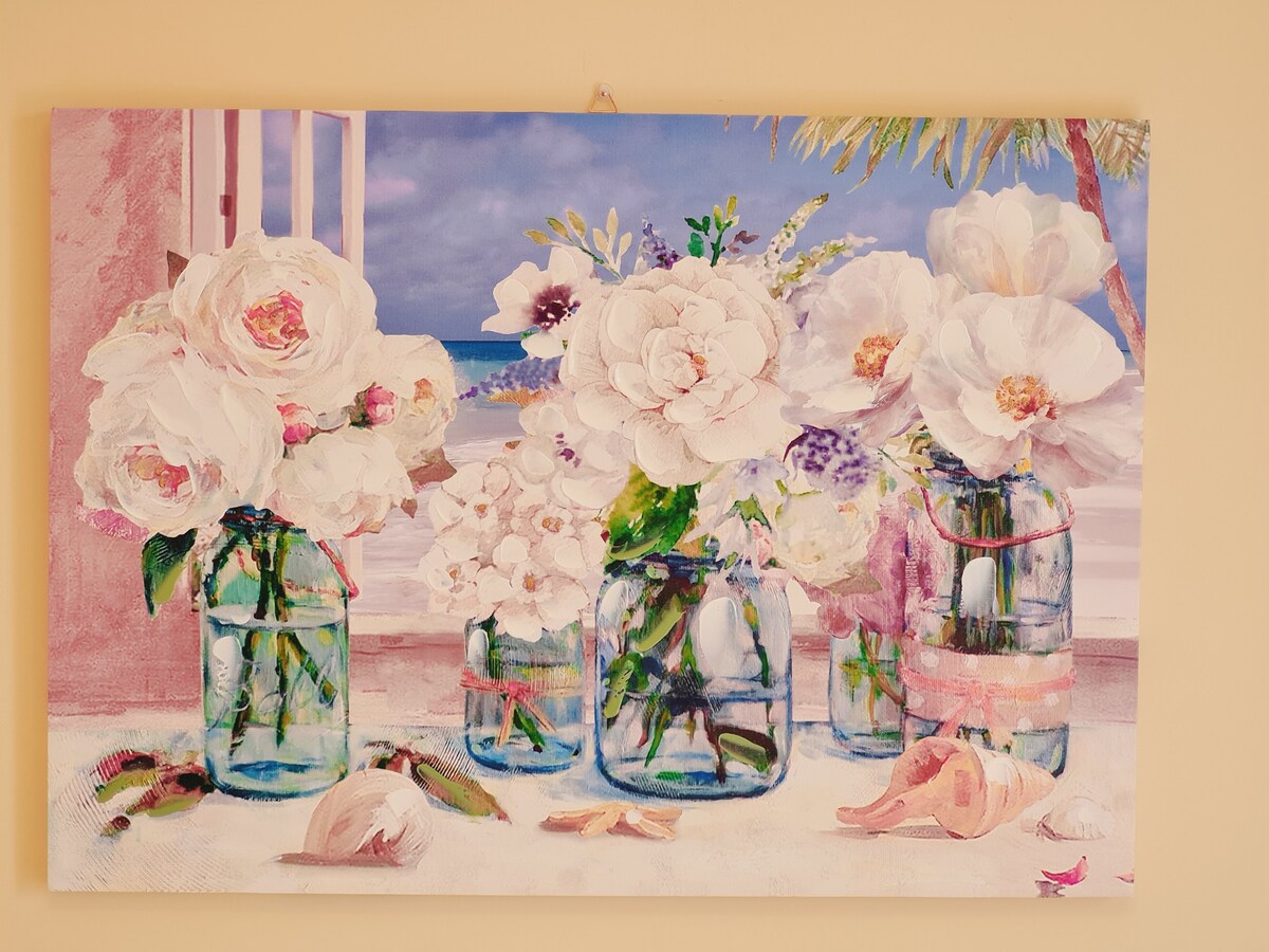 A colorful artwork displays arrangements of white flowers in various glass jars, positioned against a light background. The composition features soft petals and shells scattered on a flat surface, with hints of a beach scene visible in the background.