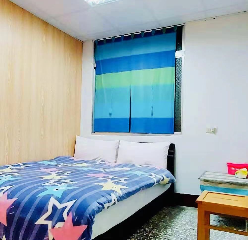 A comfortable sleeping area is presented with a spacious bed covered in a star-patterned blanket. The bed is flanked by light-colored walls and a window dressed in blue and green curtains. A wooden table sits nearby, providing additional surface space.