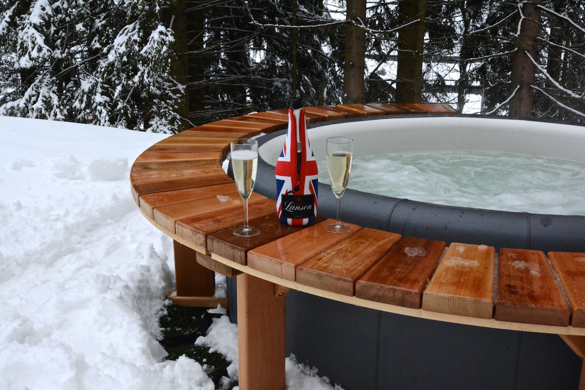 An outdoor whirlpool is surrounded by a wooden deck, with two glasses of sparkling drink set on the edge. A decorative item featuring a British flag is placed nearby. Snow-covered ground and evergreen trees create a serene winter environment.