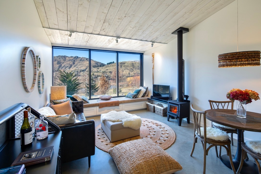 A modern lounge area features large windows showcasing scenic mountain views. A wood stove provides warmth, while a round dining table and comfortable seating invite relaxation. Natural light illuminates the room, complemented by cozy textiles and decor items that create a welcoming ambiance.