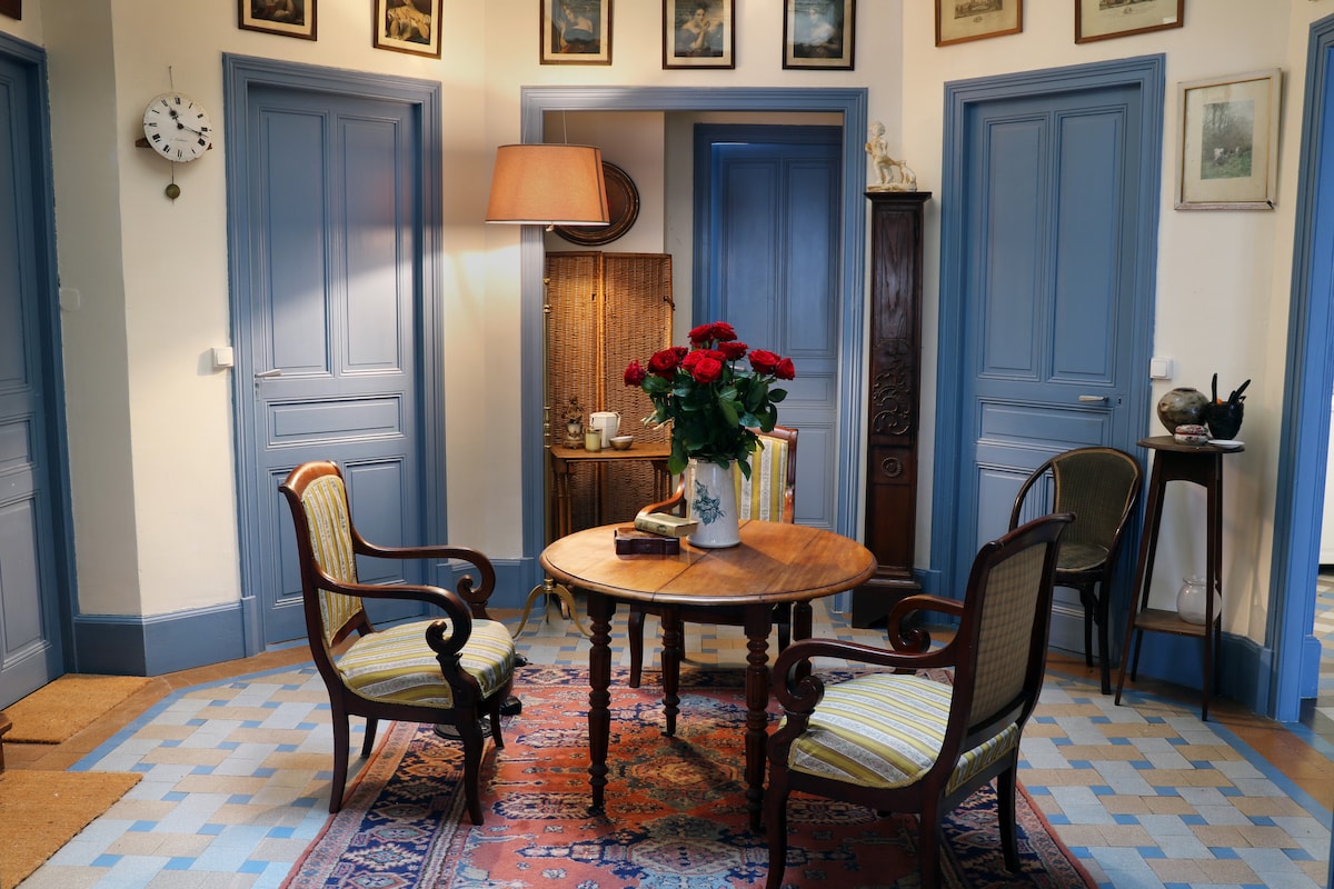 A welcoming entryway features a round wooden table at its center, surrounded by two elegantly designed chairs. A vase with fresh roses sits atop the table. Light is softly filtered through a lamp, while framed photographs adorn the walls. Muted blue doors lead to adjacent rooms.