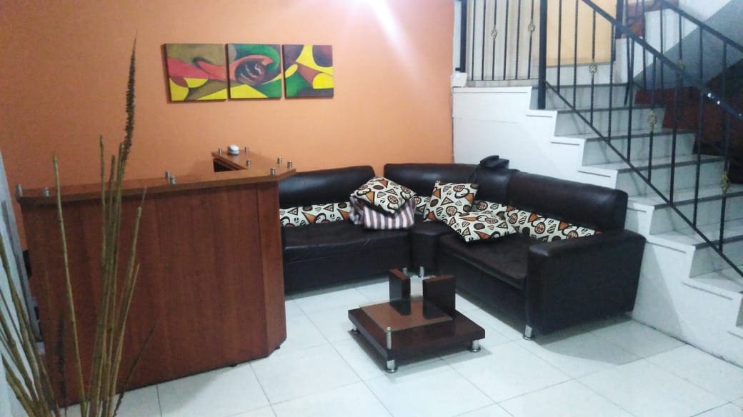 A comfortable seating area features a dark leather sectional sofa adorned with patterned cushions. A small coffee table sits in the center, complemented by a simple wooden reception desk. Colorful wall art adds a touch of personality, while a staircase is visible in the background.