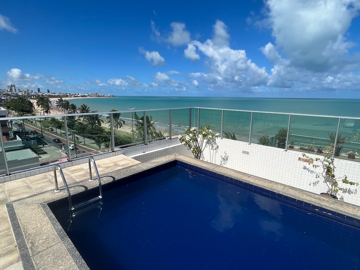 A rooftop pool is visible, surrounded by a glass railing, with clear blue water reflecting the sky. The horizon showcases the coastline of Manaíra, lined with palm trees and buildings. The scene is accented by a few potted plants near the pool.