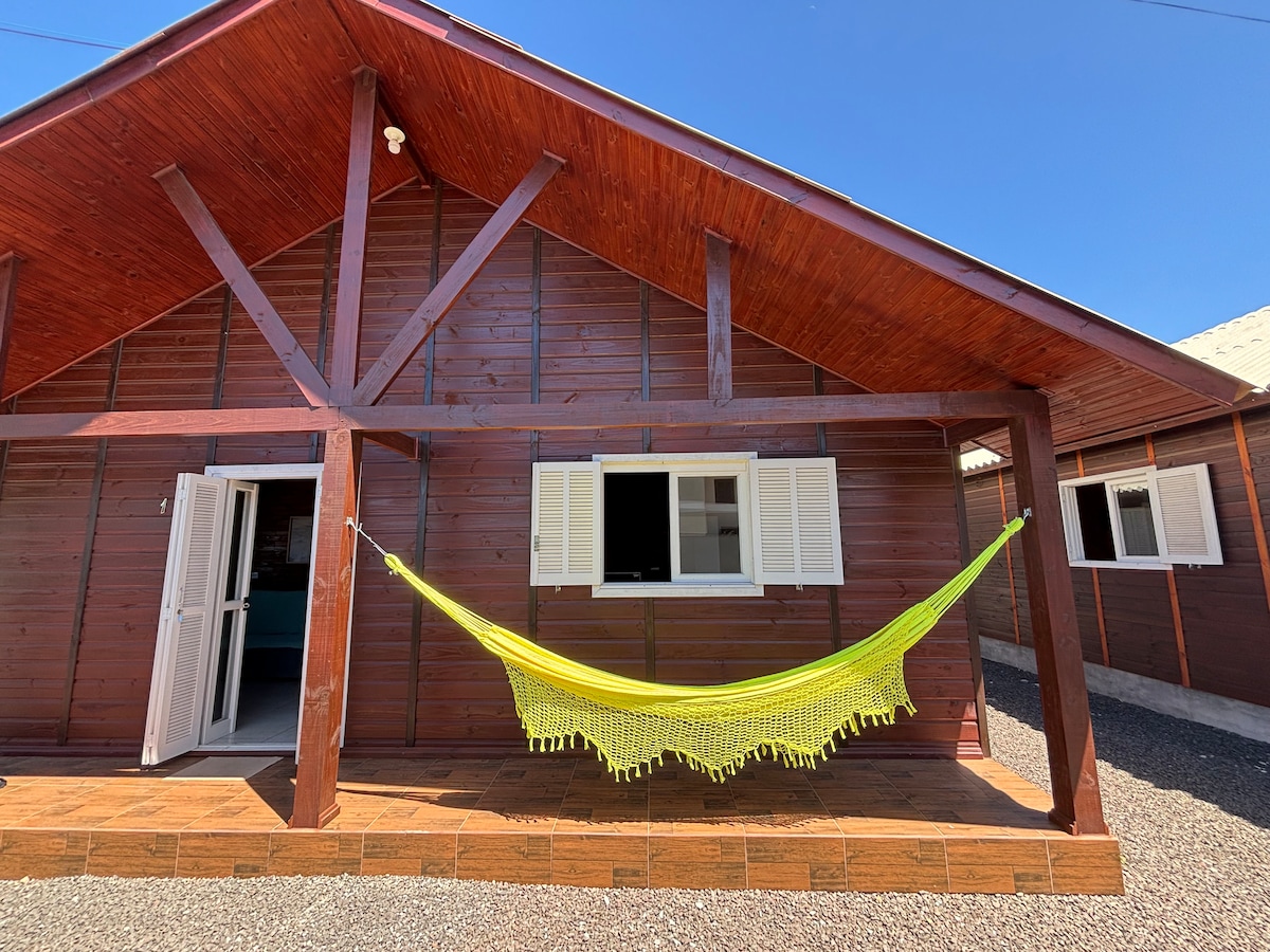 The exterior of a wooden chalet features a vibrant yellow hammock suspended between two sturdy beams. Two white windows are adorned with shutters, allowing natural light to enter the interior. The surrounding area is gravelled, and a welcoming entrance is visible.