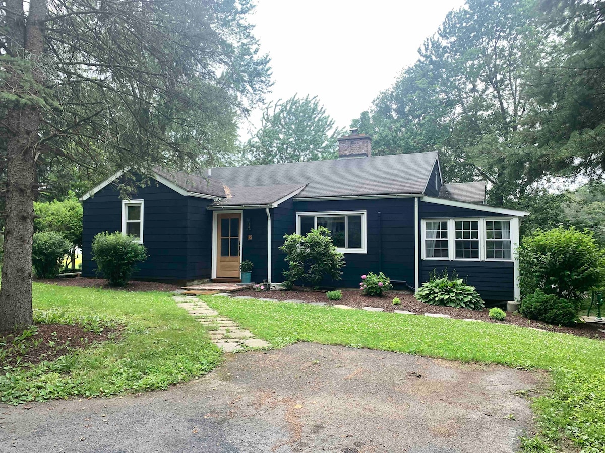 The cottage features a dark exterior with white trim and a welcoming entrance. A neat stone pathway leads to the front door, surrounded by lush greenery and well-maintained landscaping. The property is set back from the road, providing a sense of privacy.