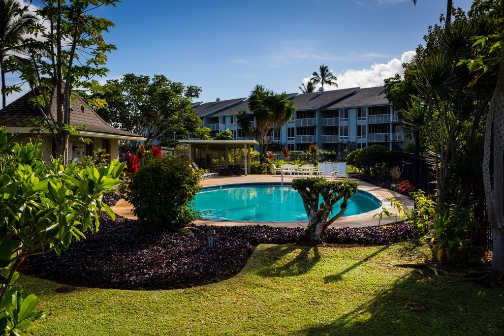 A serene outdoor pool is surrounded by lush greenery and flowering plants. The area features a well-maintained lawn and provides seating options nearby. In the background, the condo is visible with multiple windows, enhancing the inviting outdoor atmosphere.