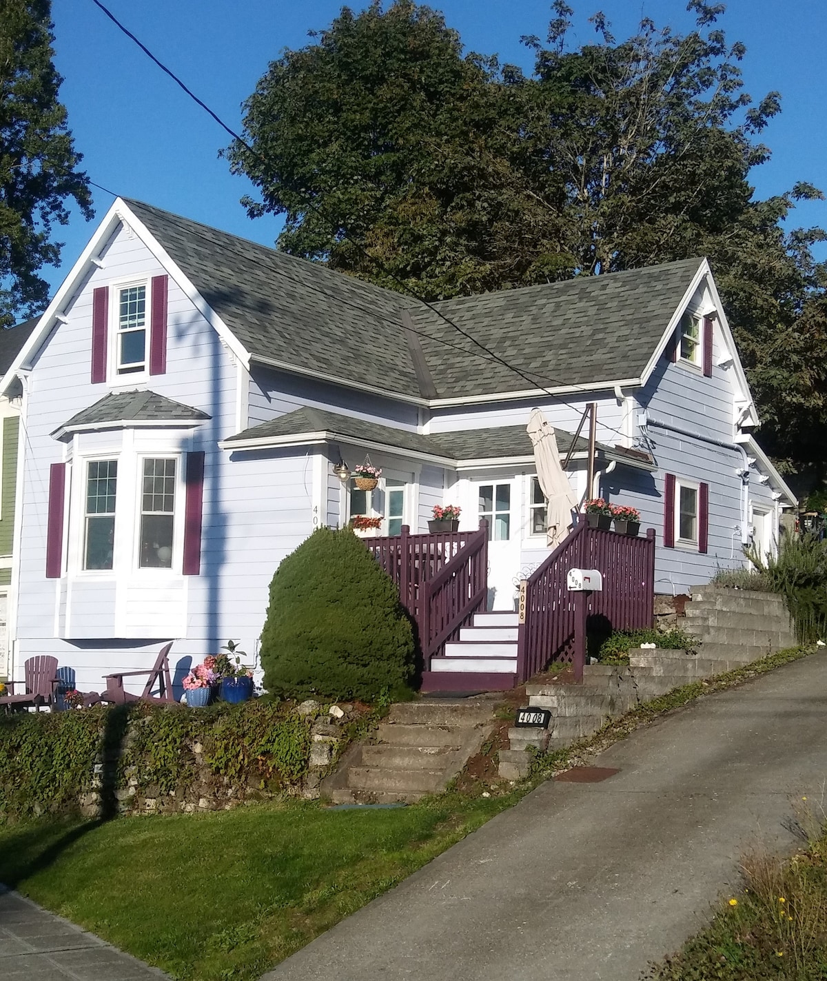 The exterior of a charming, early 20th-century house is shown, featuring pale blue siding and burgundy accents. Steps lead up to a raised porch adorned with potted plants. A well-maintained garden and trees surround the property, contributing to the serene neighborhood atmosphere.