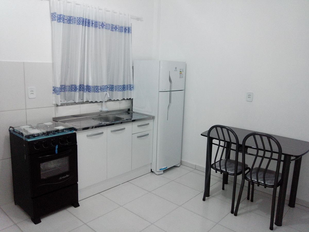 The kitchen area features a refrigerator, stove, and sink, along with a dining table that seats four. Large windows are adorned with white curtains, allowing natural light to fill the space. The cabinetry is clean and functional, providing ample storage.