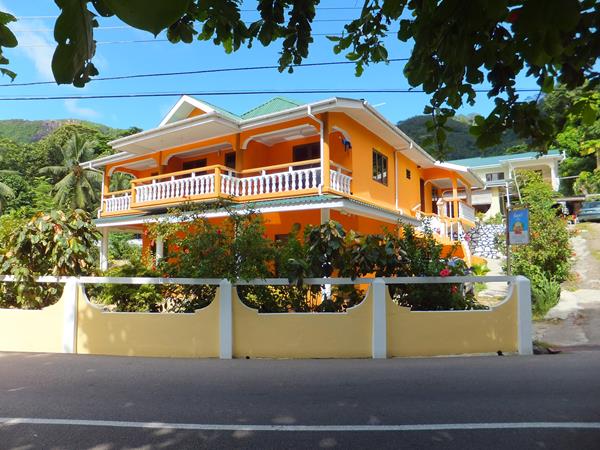 A colorful two-story building is framed by lush greenery and flowering plants. The exterior features a vibrant orange façade with white railings on the balconies. A white wall encloses the property, providing privacy from the street. The surrounding trees offer shade and a tropical ambiance.