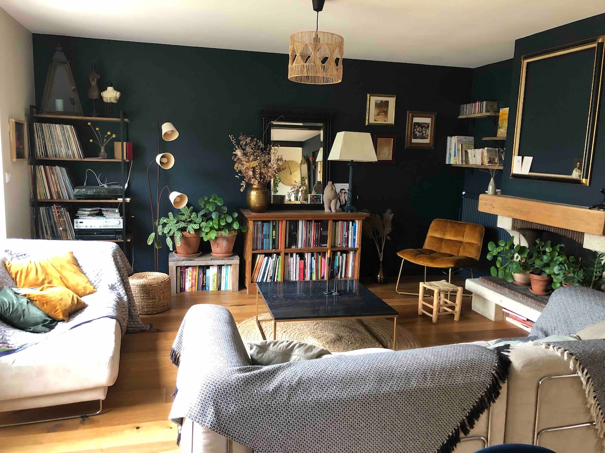 A spacious living room features a dark green accent wall complemented by natural wood flooring. A collection of houseplants adorns shelves, and a comfortable seating arrangement includes a light sofa with colorful cushions and a vintage armchair. A coffee table sits in the center, surrounded by bookshelves.