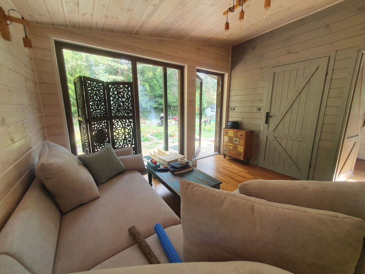 A light-filled living area features a comfortable seating arrangement with neutral-toned cushions. A patterned screen adds a decorative touch, while large glass doors offer views of the surrounding greenery. A wooden side table holds books, contributing to a relaxed atmosphere.