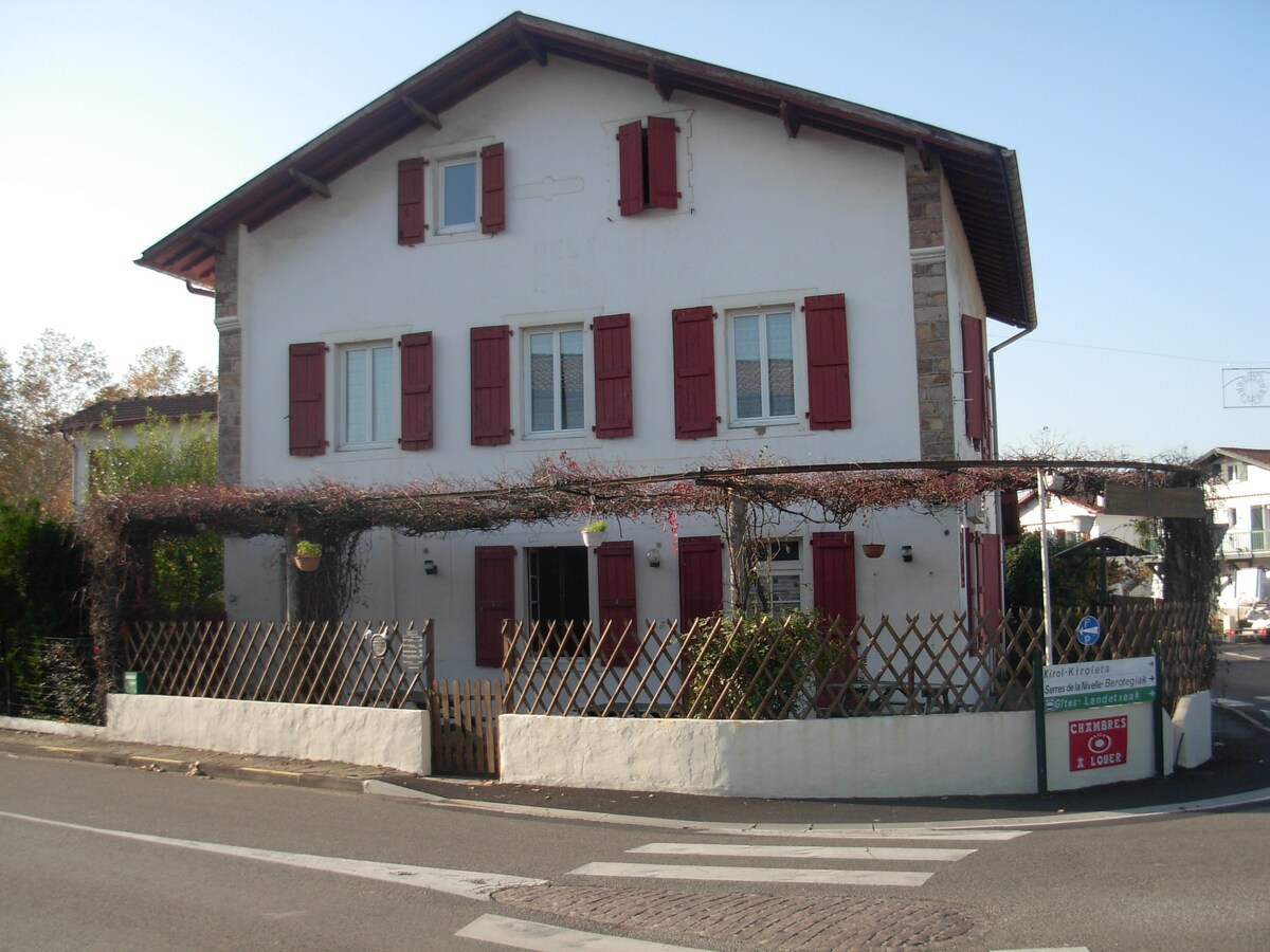 The exterior of a two-story Basque house features a combination of white walls and natural stone accents. Red shutters frame the windows, contributing to the traditional style. A wooden trellis adorned with climbing plants extends over the entrance, adding greenery to the facade.