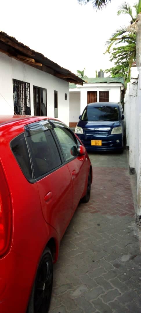 A narrow driveway is seen between two buildings, flanked by white walls. A red car is parked in the foreground, while a blue vehicle is positioned further back. The surrounding area features a mix of greenery and hard surfaces.