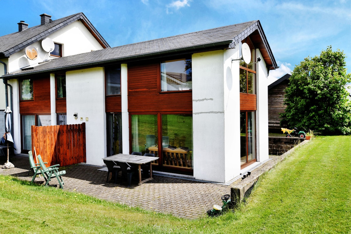 A two-story house with a sleek design features large floor-to-ceiling windows that invite ample natural light. The outdoor area is characterized by a patio table surrounded by green grass and outdoor seating. The surrounding landscape includes trees and an open view of the horizon.