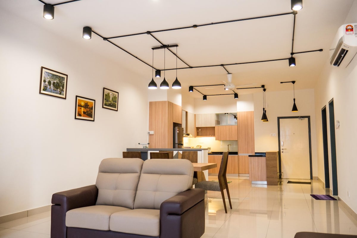 A modern living area is observed with a neutral color palette, featuring a comfortable seating arrangement and a dining table. The kitchenette showcases wooden cabinetry and built-in appliances, while pendant lights provide illumination. Wall art enhances the ambiance, and natural light brightens the space.